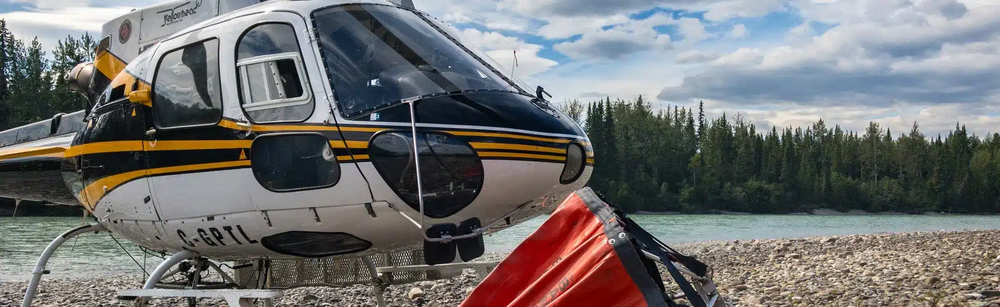 AS350B2 helicopter parked next on a riverbank during BC Wildfire service, bambi bucket nearby